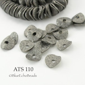May include: Close-up of gunmetal-colored, textured metal beads. The beads are teardrop-shaped with a hole in the center. Some beads are scattered, while others are in a coiled strand. The text "ATS 110" and "©BlueEchoBeads" are visible.