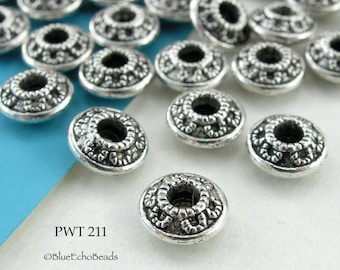 May include: A close-up of a pile of silver-toned metal beads with an intricate design. The beads are round with a hole in the center and have a raised, ornate pattern. The beads are scattered on a blue and white surface. The text "PWT 211 ©BlueEchoBeads" is visible in the lower left corner of the image.