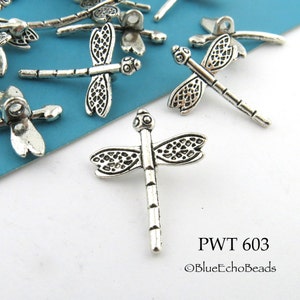 May include: Silver-toned dragonfly charms with intricate details. The charms have a loop at the top for attaching to jewelry or other crafts. The charms are scattered on a blue surface. PWT 603 ©BlueEchoBeads
