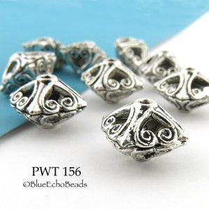 May include: A close-up of silver metal beads with a decorative design. The beads are shaped like a square with a pointed top. The beads are arranged in a scattered pattern on a white surface.