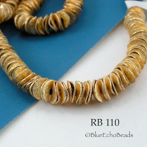 May include: A strand of gold-colored metal beads, each bead is shaped like a crescent moon. The beads are arranged in a circular pattern. The text "RB 110 ©BlueEchoBeads" is visible below the beads.