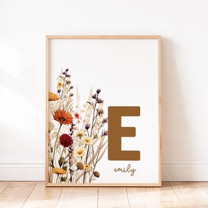 May include: A framed art print with a floral design in shades of orange, yellow, and purple. A large brown letter "E" is centered, with the name "emily" below in script. The artwork is set against a white background.
