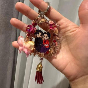 以下が含まれることがあります： フォーマルな服装の二人のキャラクターが描かれた円形のフレームが付いた装飾的なキーホルダー。フレームにはピンクと赤の花のアクセント、半透明のピンクの蝶のチャーム、そして濃い赤の布製タッセルが飾られています。