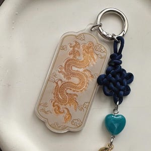 May include: A clear acrylic keyring featuring a gold dragon design and a silver ring. It includes a navy blue knot and a turquoise heart charm. The dragon is set against a background of cloud motifs.