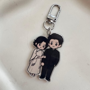 Peut inclure: Un porte-clés en acrylique transparent représentant une illustration de deux personnages côte à côte. L'un porte une robe couleur crème à col montant, l'autre un costume noir. Le porte-clés est doté d'un fermoir en métal argenté.