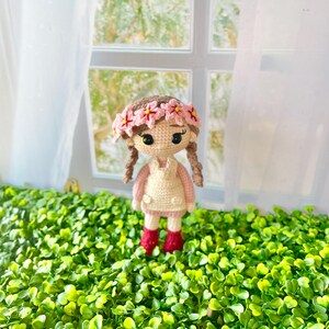 May include: A crocheted doll with brown hair wearing a pink flower crown, a pink and white dress, and red boots. The doll is standing on a bed of green artificial grass.