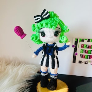 Puede incluir: Muñeca de crochet verde con ropa a rayas blancas y negras, botas negras y un globo rosa. La muñeca tiene grandes ojos negros y cabello verde. La muñeca está de pie sobre una base de madera.