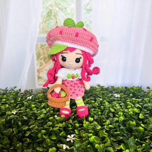 Puede incluir: Muñeca de ganchillo hecha a mano que representa a una niña fresa. La muñeca tiene pelo rosa, un sombrero de fresa rosa y un vestido rosa con lunares blancos. Sostiene una pequeña cesta con fresas en miniatura. La muñeca está sobre follaje verde.