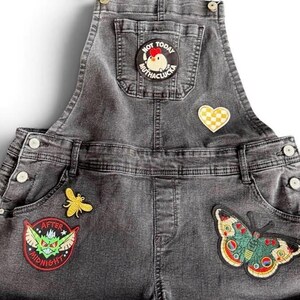 May include: Dark gray denim overalls featuring a chest pocket with a circular patch showing a chicken and the text NOT TODAY MUTHACLUCKA. Additional patches include a yellow checkered heart, a yellow bee, a green creature, and a colorful butterfly.