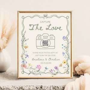 Könnte beinhalten: Gerahmtes Hochzeitsschild mit dem Text "Capture The Love" und einer Kamera-Grafik. Das Schild enthält einen QR-Code und die Namen "Anastasia & Christian" mit dem Datum 02.14.2030. Blumige Akzente und ein goldener Rahmen runden das Design ab.