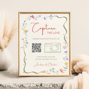 May include: A framed sign with the words "Capture the Love" in red script, surrounded by floral accents. The sign includes a QR code and a camera illustration, inviting guests to share photos. The names "Anastasia & Christian" are at the bottom.