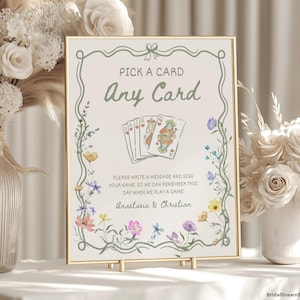 Puede incluir: Un letrero enmarcado con el texto "PICK A CARD Any Card" e ilustraciones florales. El letrero incluye el texto "PLEASE WRITE A MESSAGE AND SIGN YOUR NAME: SO WE CAN REMEMBER THIS DAY WHEN WE PLAY A GAME! Anastasia & Christian."