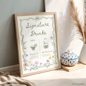 May include: A framed sign with the words "Signature Drinks" in elegant script. The sign features floral accents and drink illustrations for "Hers" and "His", including "French 75" and "Old Fashioned".
