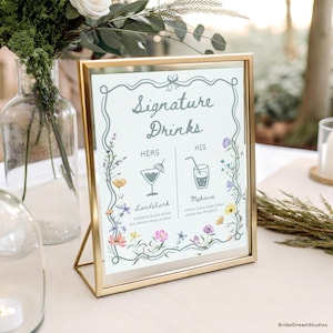 May include: A gold-framed sign with the words "Signature Drinks" in elegant script. The sign features floral illustrations and details two drink options: "Landshark" and "Mykonos". The sign is displayed on a white tablecloth.