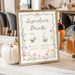May include: A framed sign with the text "Signature Drinks" and floral illustrations. The sign lists "French 75" and "Old Fashioned" cocktails. A white pumpkin and bottles are in the background.