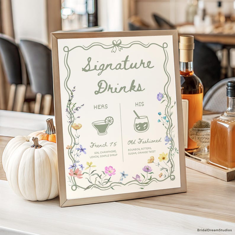 May include: A framed sign with the text "Signature Drinks" and floral illustrations. The sign lists "French 75" and "Old Fashioned" cocktails. A white pumpkin and bottles are in the background.