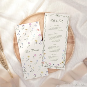 Peut inclure: Deux cartons de menu de mariage avec des motifs floraux. L'un des cartons indique "The Menu" et l'autre "Let's Eat" avec une liste d'éléments de menu. Les cartons sont sur un plateau en bois.