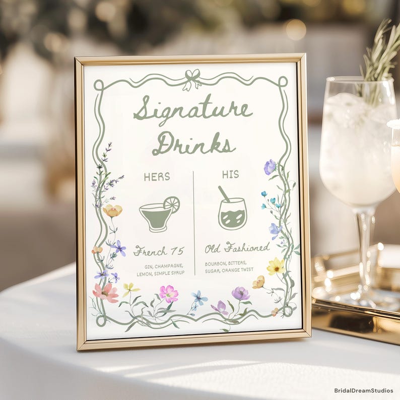 May include: A framed sign with the words "Signature Drinks" in elegant script. The sign features floral illustrations and lists "French 75" and "Old Fashioned" cocktails. The sign is in a gold frame and is set on a white surface.