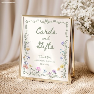 May include: A gold-framed sign with the words "Cards and Gifts" in green script, surrounded by a floral border. The sign also includes the text "Thank You" and names and a date. The sign is displayed on a woven surface.
