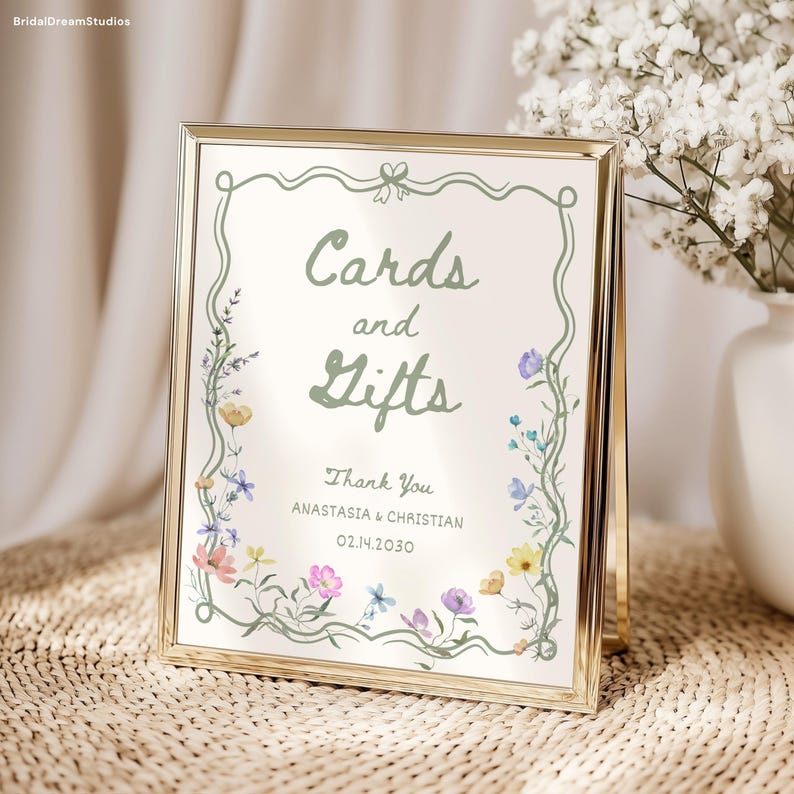May include: A gold-framed sign with the words "Cards and Gifts" in green script, surrounded by a floral border. The sign also includes the text "Thank You" and names and a date. The sign is displayed on a woven surface.