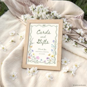 May include: A wooden framed sign with the words "Cards and Gifts" in green script, surrounded by a floral border. The sign also includes the names "Anastasia & Christian" and the date "02.14.2020". White flowers are scattered around the sign.