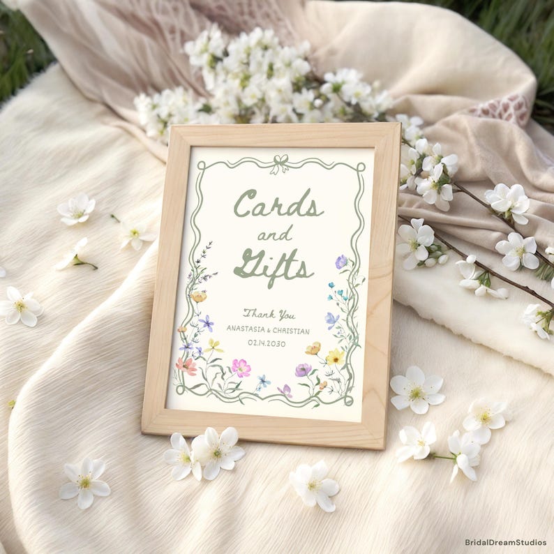 May include: A wooden framed sign with the words "Cards and Gifts" in green script, surrounded by a floral border. The sign also includes the names "Anastasia & Christian" and the date "02.14.2020". White flowers are scattered around the sign.