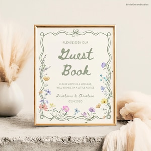 Puede incluir: Un letrero enmarcado con las palabras "Guest Book" en una fuente decorativa. El letrero está adornado con ilustraciones florales en colores pastel e incluye los nombres "Anastasia & Christian" y la fecha 02.14.2030.
