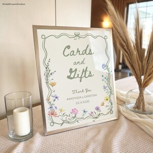 May include: A framed sign with the words "Cards and Gifts" in green script, surrounded by a floral border. The sign also includes the text "Thank You" and names and a date. A glass candle holder and vase with dried flowers are also visible.