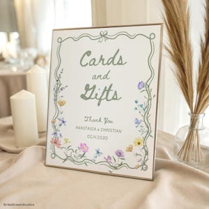 May include: A rectangular sign with the words "Cards and Gifts" in green script, framed by a floral border. The sign includes the text "Thank You" and names, with a date. The background is off-white, and the frame is a light brown.
