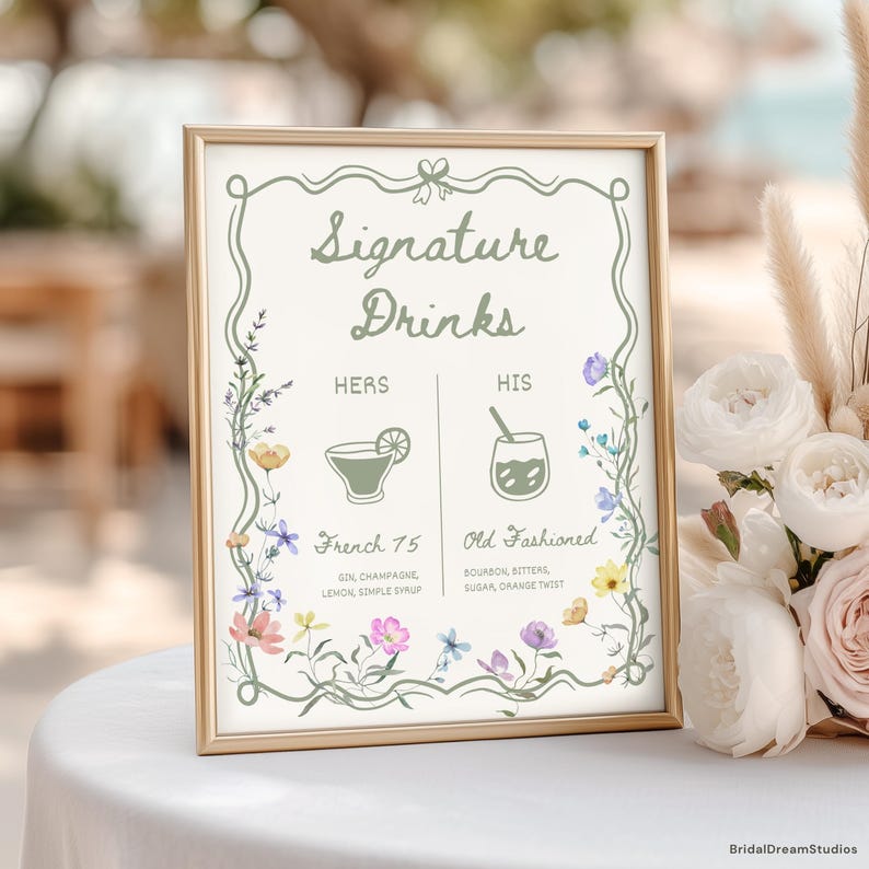 May include: A framed sign with the words "Signature Drinks" in elegant script. The sign features floral illustrations and drink names: "French 75" and "Old Fashioned." The frame is gold, and the sign is displayed on a white surface.