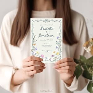 May include: A rectangular wedding invitation card with a cream background, featuring a blue decorative border and colorful wildflower illustrations. The text reads: Join us for the wedding of Isabella & Jonathan, November 19th, 2031, The Groove, 8080 Al Garrison St, Las Vegas, Nevada.