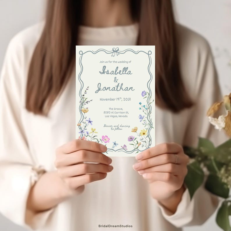 May include: A rectangular wedding invitation card with a cream background, featuring a blue decorative border and colorful wildflower illustrations. The text reads: Join us for the wedding of Isabella & Jonathan, November 19th, 2031, The Groove, 8080 Al Garrison St, Las Vegas, Nevada.