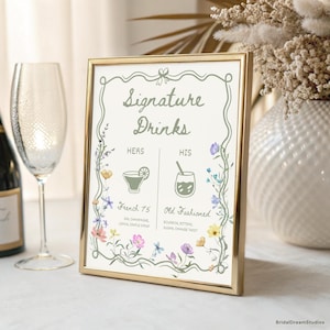 May include: A framed sign with the words "Signature Drinks" in elegant script. The sign features illustrations of a French 75 and an Old Fashioned cocktail, with floral accents. The frame is gold, and the background is off-white.