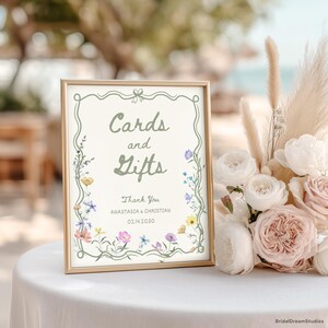May include: A framed sign with the words "Cards and Gifts" in green script, surrounded by floral illustrations. The sign also includes the names "Anastasia & Christian" and the date "02.14.2030". A bouquet of white and pink roses is next to the sign.