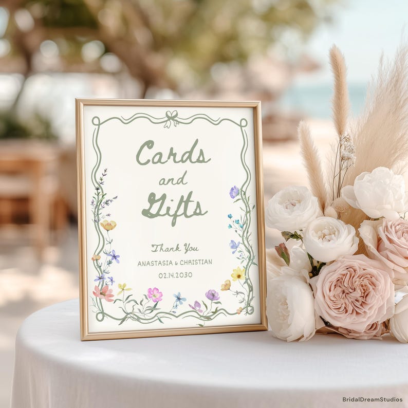 May include: A framed sign with the words "Cards and Gifts" in green script, surrounded by floral illustrations. The sign also includes the names "Anastasia & Christian" and the date "02.14.2030". A bouquet of white and pink roses is next to the sign.