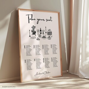 May include: A framed wedding seating plan with a light background and a wooden frame. The top of the chart reads "Take your seat" in cursive. The chart includes illustrations of food and drink, and guest names are listed in columns.