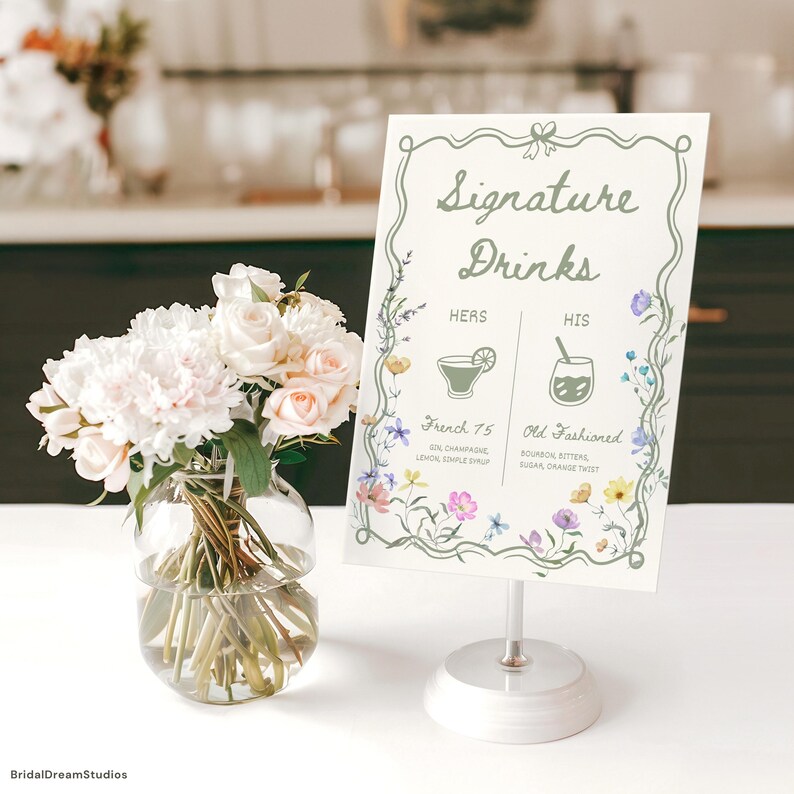 May include: A white sign with "Signature Drinks" in green script, floral accents, and drink illustrations. The sign is on a silver stand, next to a vase of flowers. The sign lists "French 75" and "Old Fashioned" cocktails.