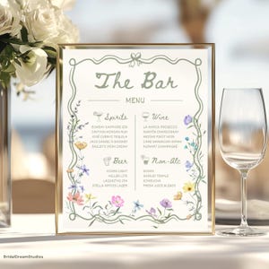 以下が含まれることがあります： 「The Bar」の文字と花柄が施された額入りのバーメニュー。メニューには、スピリッツ、ワイン、ビール、ノンアルコール飲料が記載されています。右側にはワイングラスがあります。メニューは白い表面にあります。