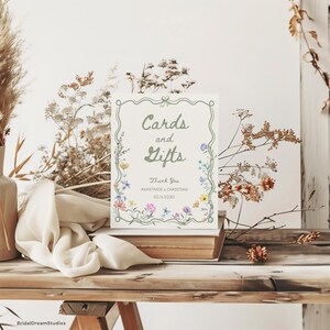 May include: A rectangular sign with the words "Cards and Gifts" in a decorative font, framed by a floral border. The sign also includes the text "Thank You, Anastasia & Christian 02.14.2030". The sign is placed on a stack of books on a wooden table.