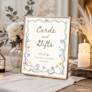 May include: A framed sign with the words "Cards and Gifts" in green script, surrounded by a floral border. The sign includes the names Anastasia & Christian and the date 02.14.2030. The frame is gold.
