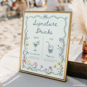 May include: A framed sign with gold trim displays "Signature Drinks" in elegant script. The sign features floral accents and drink illustrations, with "Hers" and "His" drink options listed below. The "Landshark" and "Mykonos" drink recipes are also included.