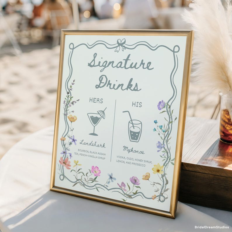May include: A framed sign with gold trim displays "Signature Drinks" in elegant script. The sign features floral accents and drink illustrations, with "Hers" and "His" drink options listed below. The "Landshark" and "Mykonos" drink recipes are also included.