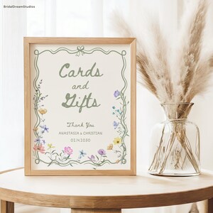 May include: A framed sign with the words "Cards and Gifts" in a decorative font, surrounded by floral illustrations. The sign also includes the names "Anastasia & Christian" and the date 02.14.2030. A vase with dried flowers sits beside the sign.
