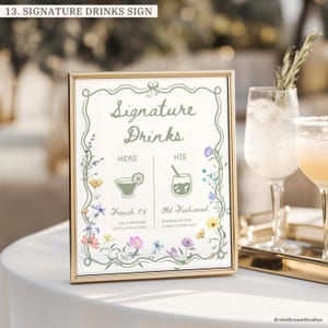 May include: A rectangular gold-framed sign titled Signature Drinks with a green border and floral illustrations. It lists French 75 under Hers and Old Fashioned under His, including ingredients for each cocktail. The sign is displayed on a white surface.
