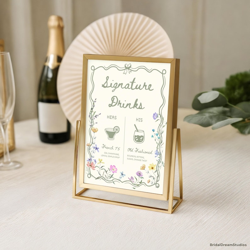 May include: A gold metal framed sign with the words "Signature Drinks" in elegant script. The sign features floral illustrations and drink details for "Hers" and "His" cocktails. A champagne bottle and fan-shaped decoration are in the background.