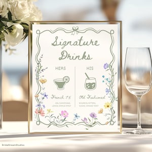 May include: A framed sign with the words "Signature Drinks" in elegant script. The sign lists "Hers" and "His" cocktails: French 75 and Old Fashioned, with ingredients. Floral accents and a gold frame complete the design. A wine glass is to the right.