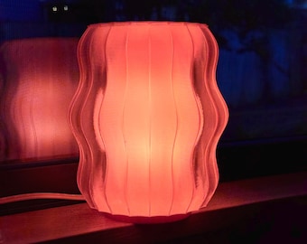 May include: A glowing, wavy-edged lamp in a soft coral color. The lamp has vertical ridges and sits on a wooden surface. The background is dark, with a hint of a window and foliage.
