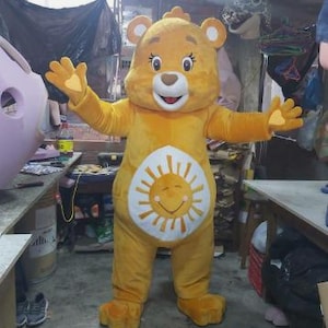 Könnte beinhalten: Ein gelbes Care Bear Maskottchen-Kostüm mit einer lächelnden Sonne auf dem Bauch. Der Bär trägt einen gelben Overall mit einem weißen Sonnendesign. Der Bär hat ein freundliches Gesicht mit schwarzen Augen und einer braunen Nase.