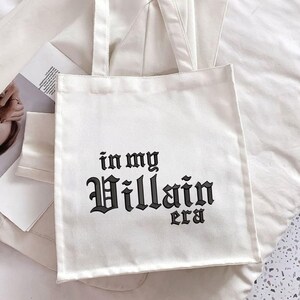 May include: White canvas tote bag with black text that reads "in my Villain era".