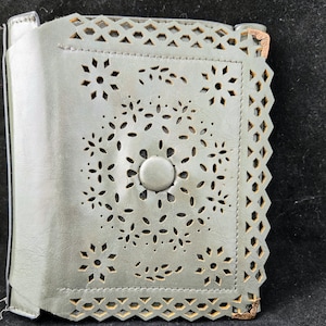 May include: A gray leather wallet with a decorative cut-out design. The wallet has a button closure and a scalloped edge.